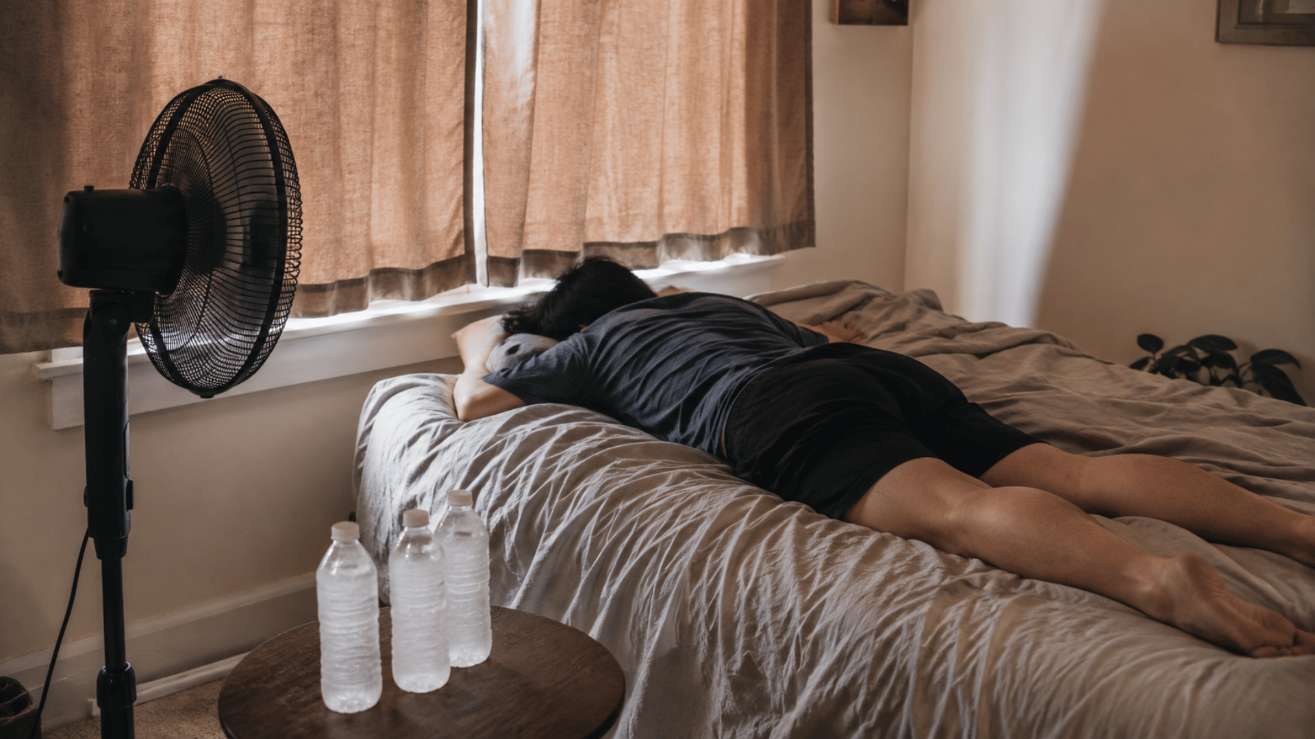 person lying on a bed with closed curtains frozen water bottles and a standing fan cooling the room