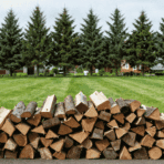 large pile of split firewood stacked on grass with evergreen trees and homes in the background