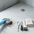 gloved hand lifting a shower drain cover beside tools on a modern tiled floor