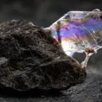 Rough mineral specimen with iridescent hues on a textured stone surface