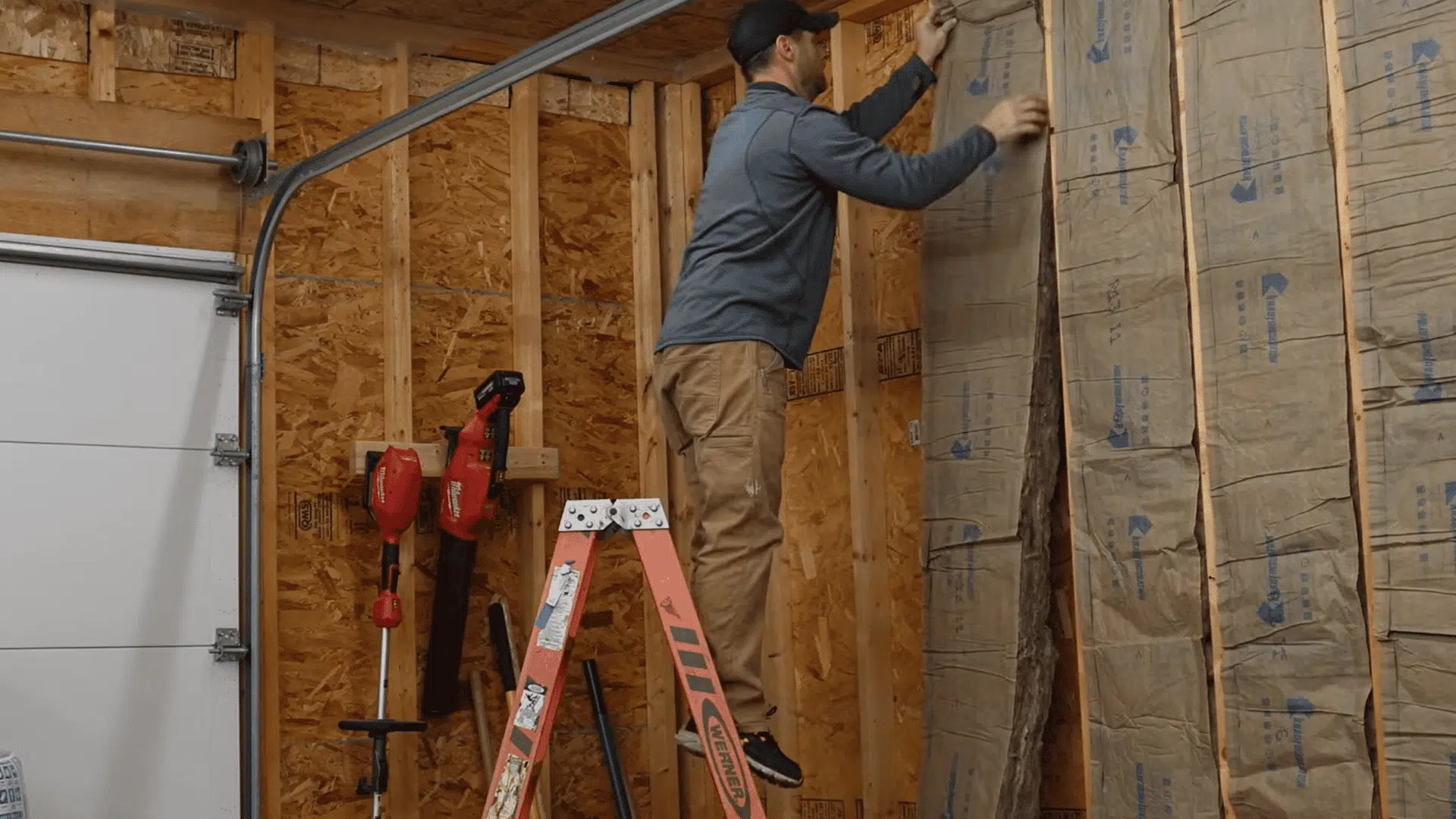 homeowner fitting fiberglass batt insulation between garage wall studs around wiring and pipes