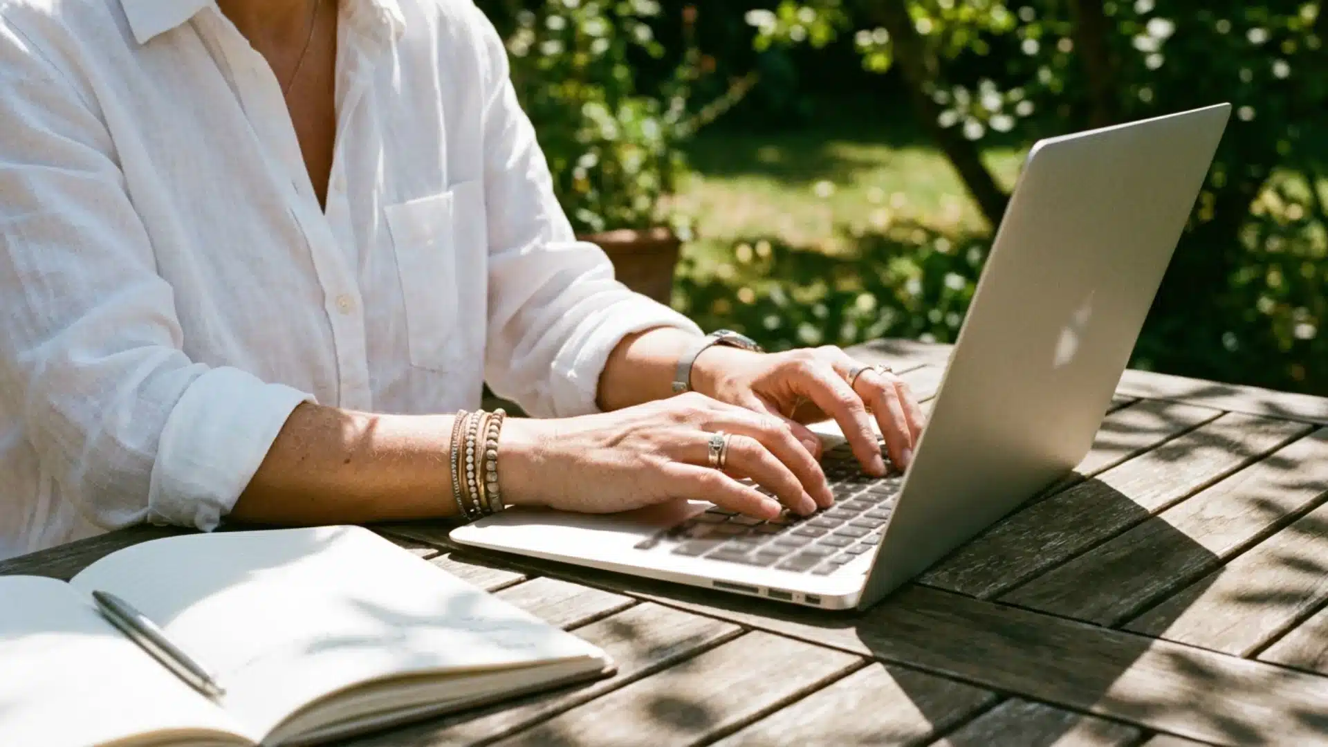 person in a white shirt types on a laptop at a sunlit outdoor table beside an open notebook
