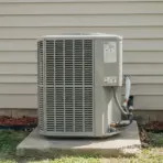 outdoor ac condenser unit on concrete pad beside beige house siding with mulch and grass in backyard