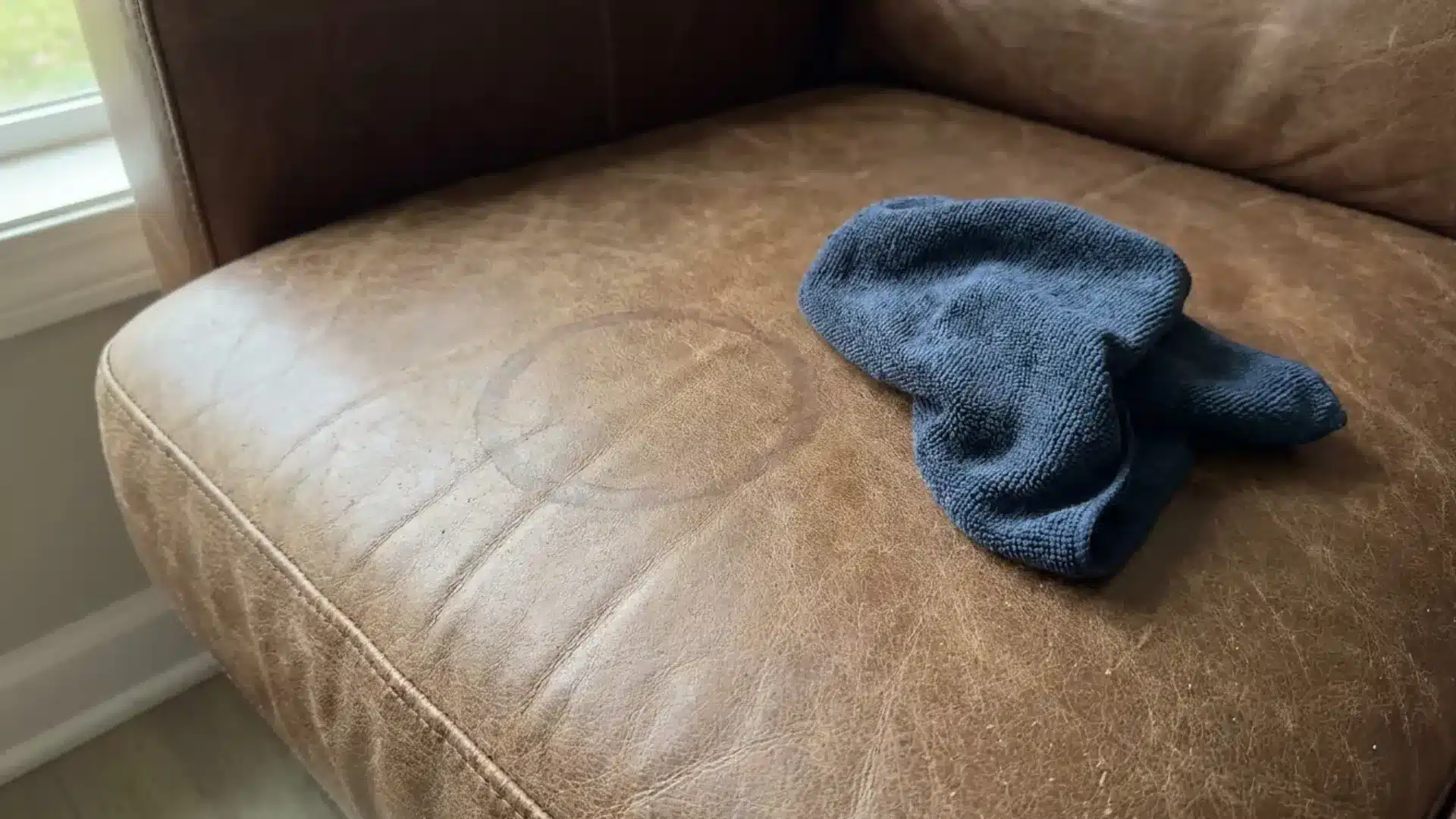 brown leather cushion with a faint water ring stain and a blue microfiber cloth beside it