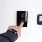 Finger pressing digital thermostat on white wall displaying temperature settings