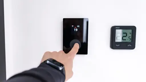 Finger pressing digital thermostat on white wall displaying temperature settings