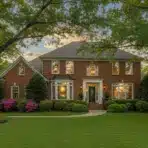 Brick colonial house with large windows and manicured lawn surrounded by tall trees and blooming bushes