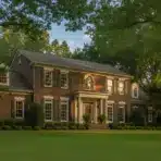 Brick colonial house with white columns and lush green lawn surrounded by tall trees