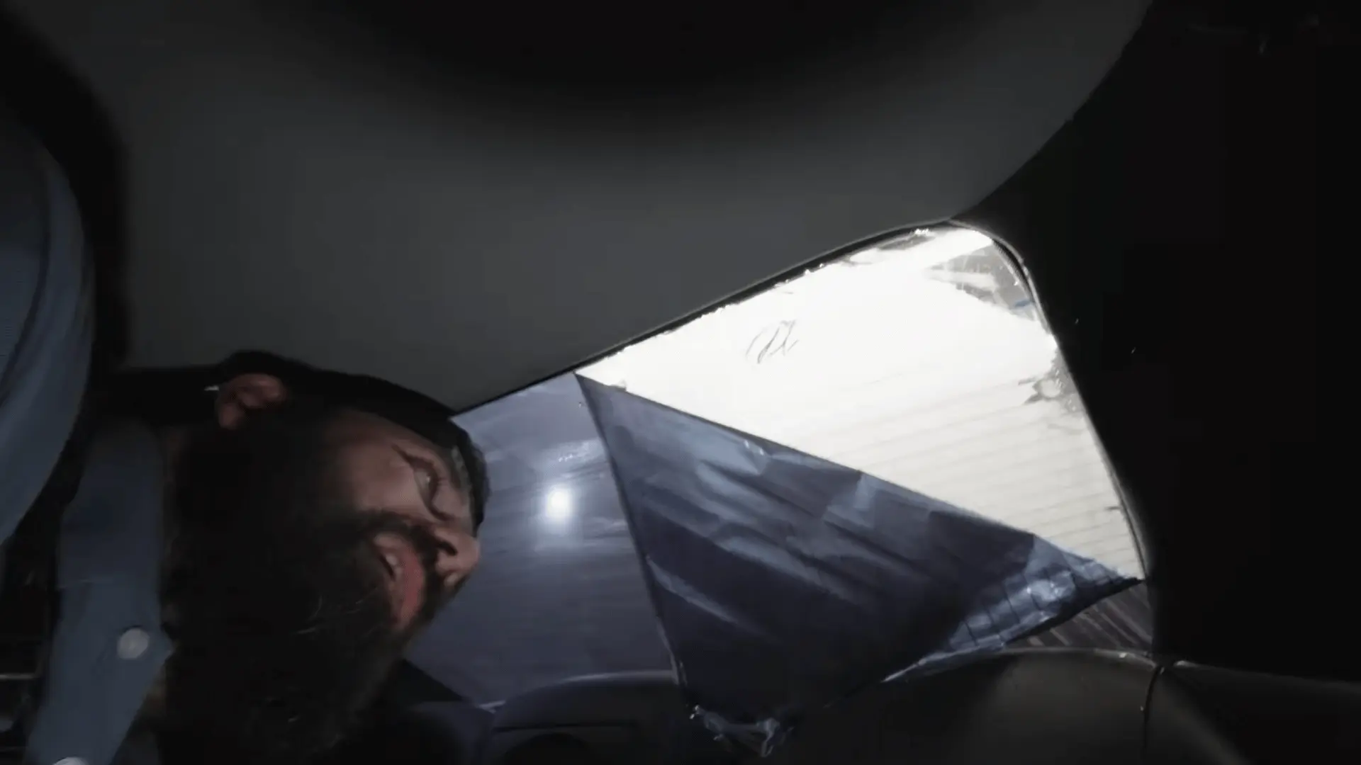 a bearded worker leans into a car while pulling dark window film across the rear windshield during installation