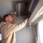 Man in beige hoodie opening overhead cabinet in bright RV interior