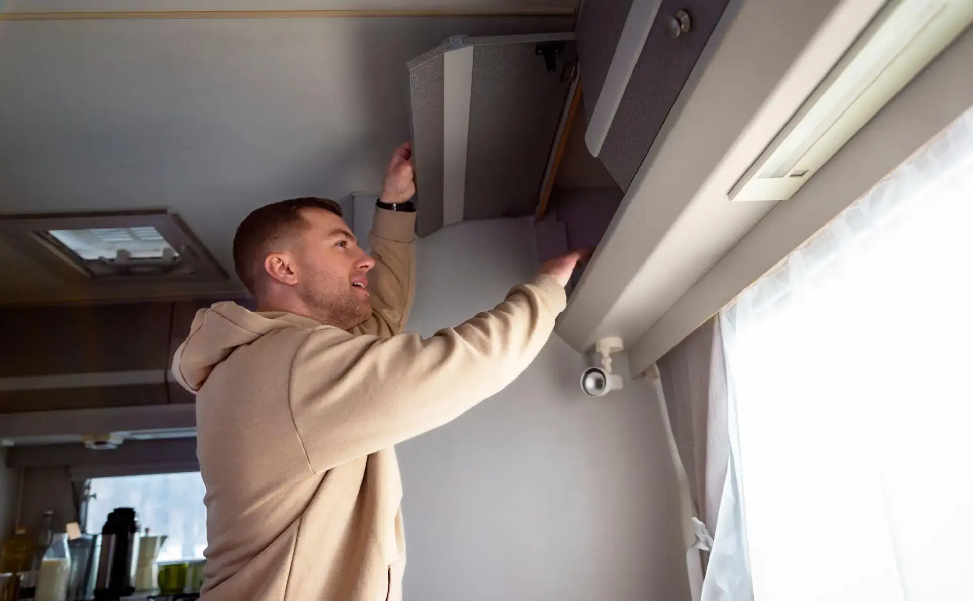 Man in beige hoodie opening overhead cabinet in bright RV interior