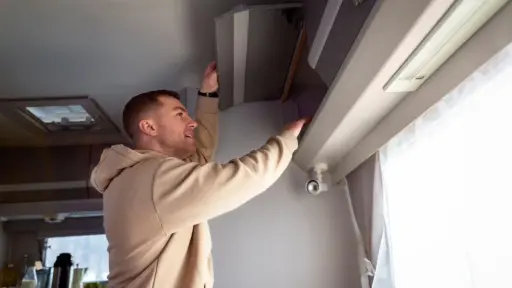 Man in beige hoodie opening overhead cabinet in bright RV interior
