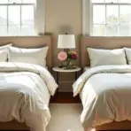 Twin beds with beige bedding in a sunlit bedroom, nightstand with flowers in between