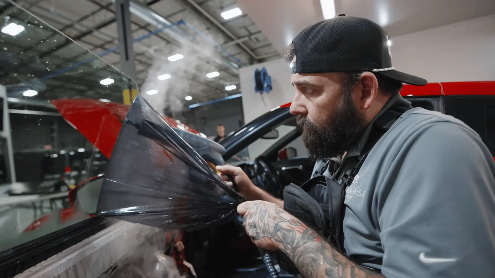 a bearded technician applies dark tint film to a raised car window while steam rises inside an automotive workshop