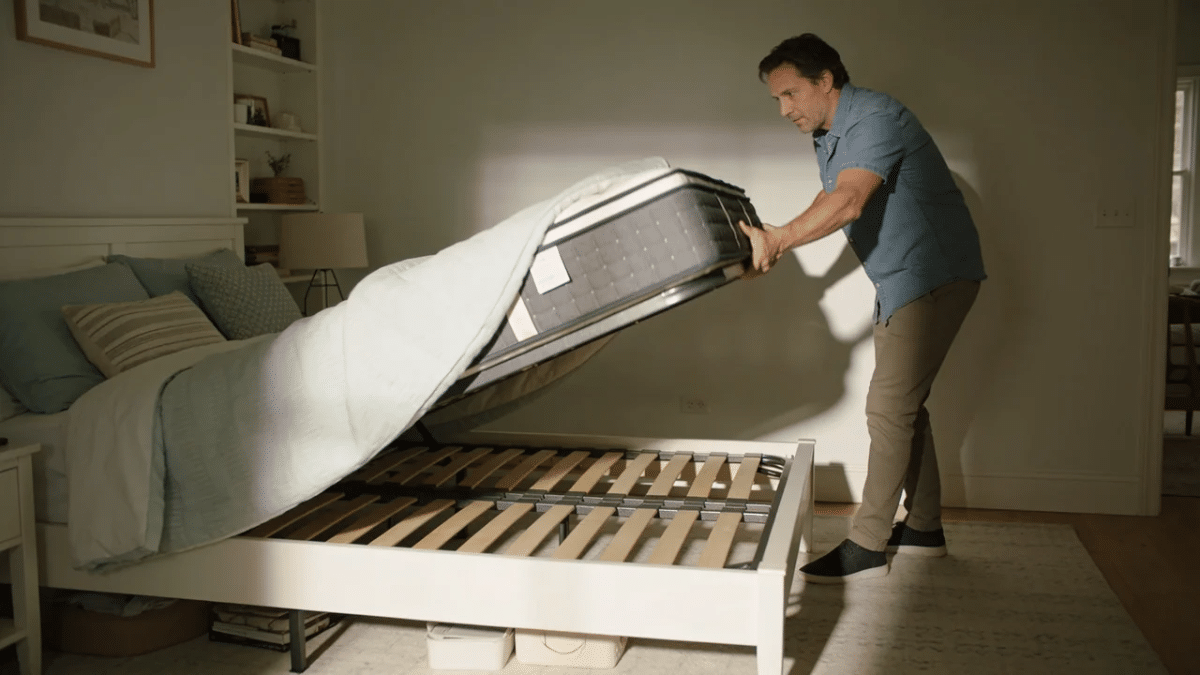homeowner rotating a mattress head to foot on a bed frame to test for squeaks in a bright bedroom