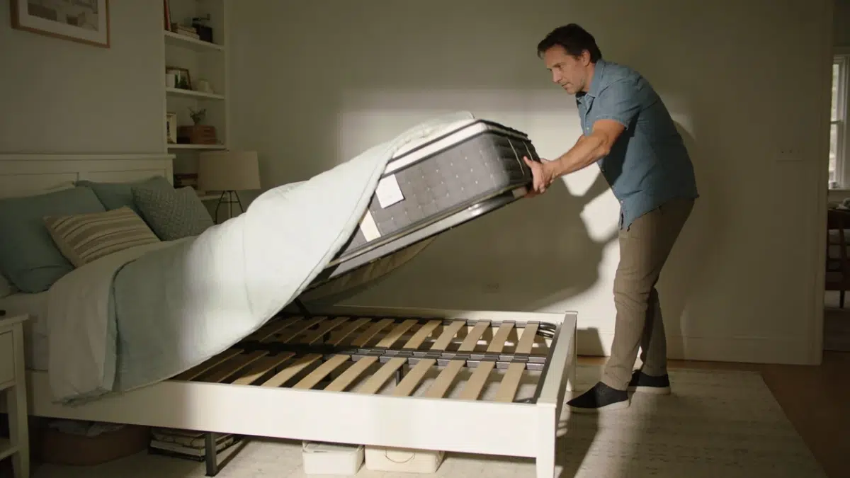 homeowner rotating a mattress head to foot on a bed frame to test for squeaks in a bright bedroom