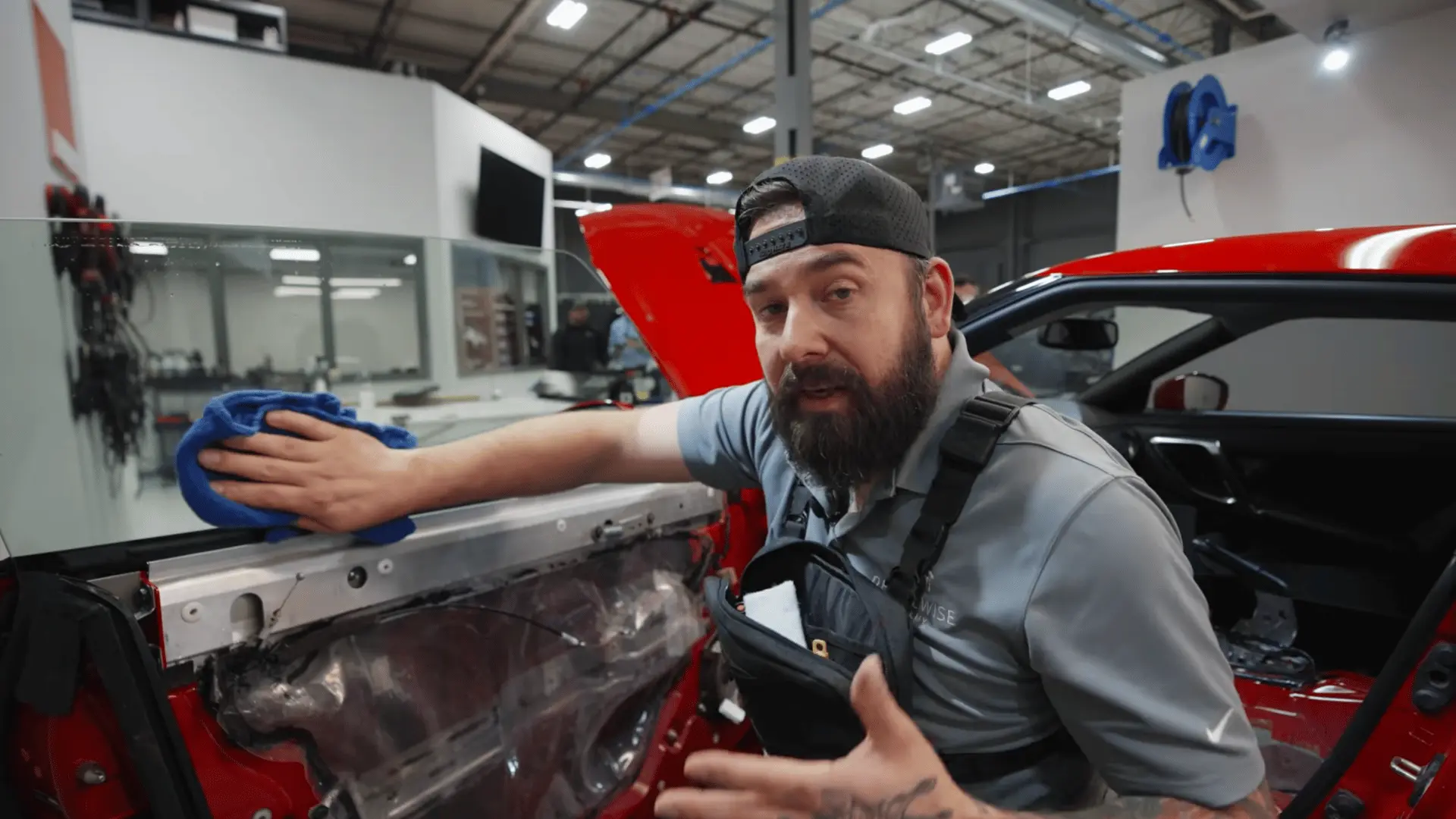 a bearded technician wipes a raised car window with a blue cloth beside an exposed red door panel in a workshop