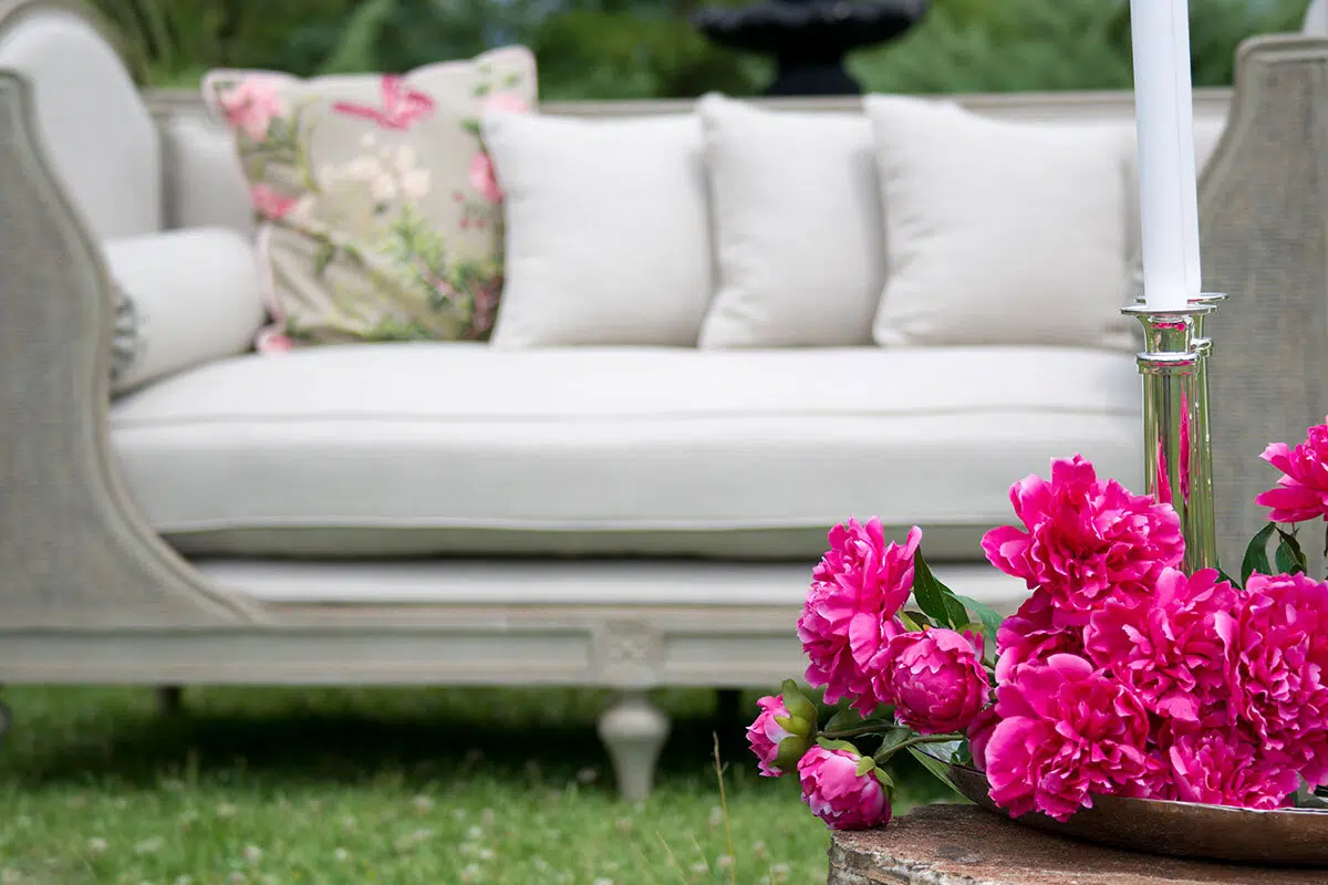 Elegant outdoor patio scene with white sofa, floral cushion, and vibrant pink peonies in foreground