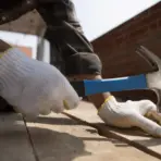 Person in white gloves hammering nail into wooden plank outdoors