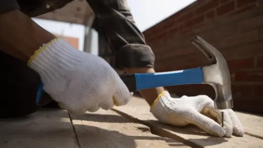 Person in white gloves hammering nail into wooden plank outdoors