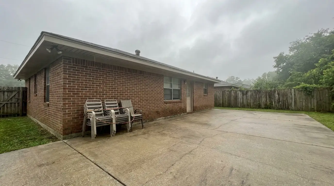 Brick house with stacked chairs on concrete patio under cloudy sky in backyard setting