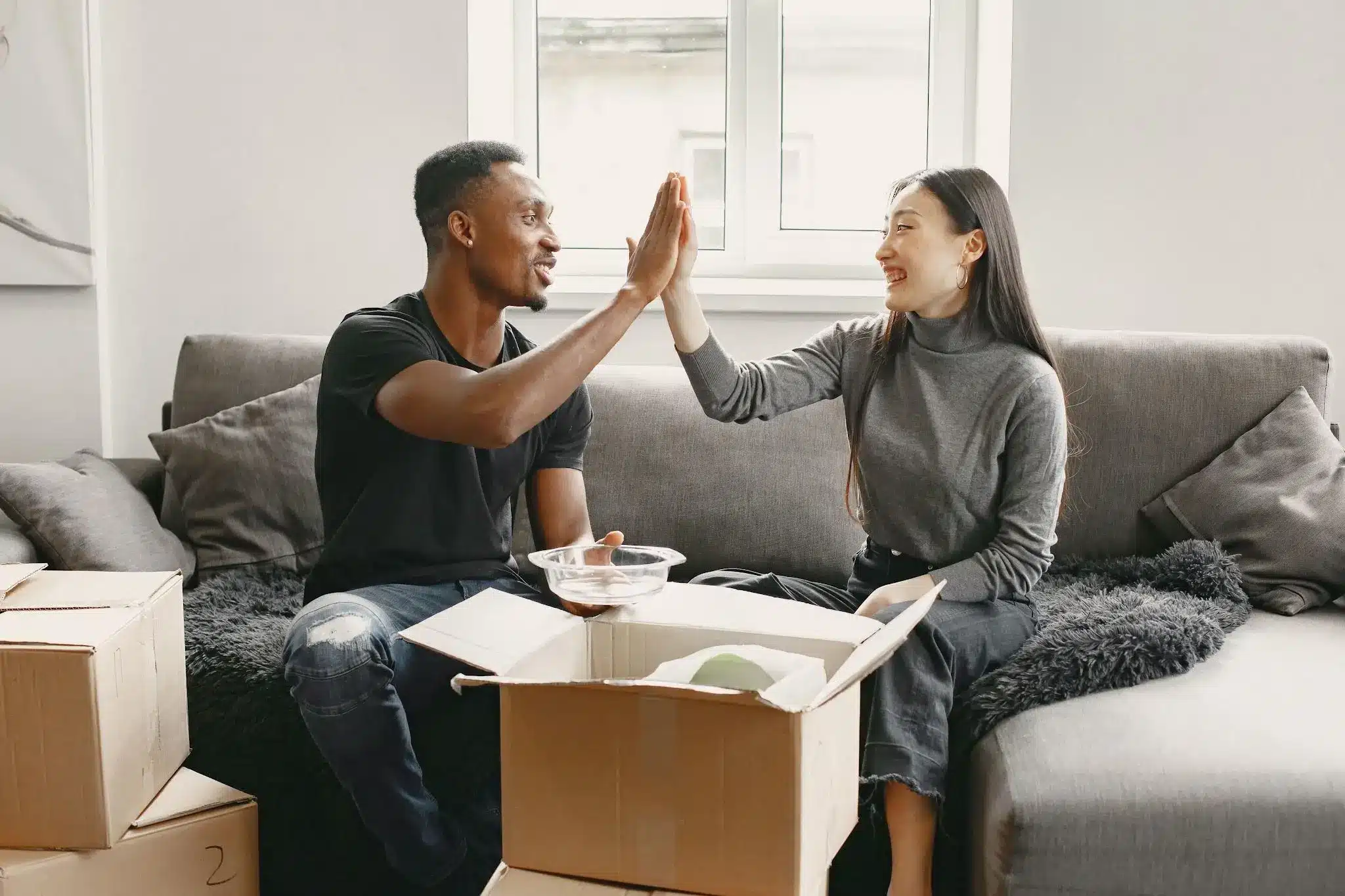 Couple high-fiving while unpacking boxes on gray sofa in bright living room
