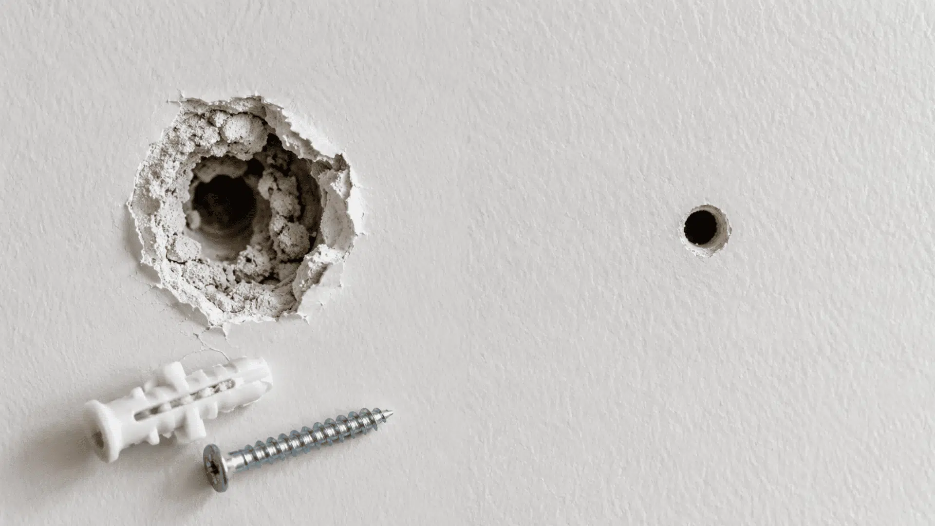 enlarged damaged drywall hole with a loose anchor beside a new mounting point in intact drywall