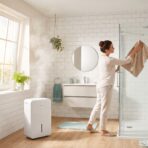 Woman cleaning glass shower door in a bright, white-tiled bathroom with plants and dehumidifier