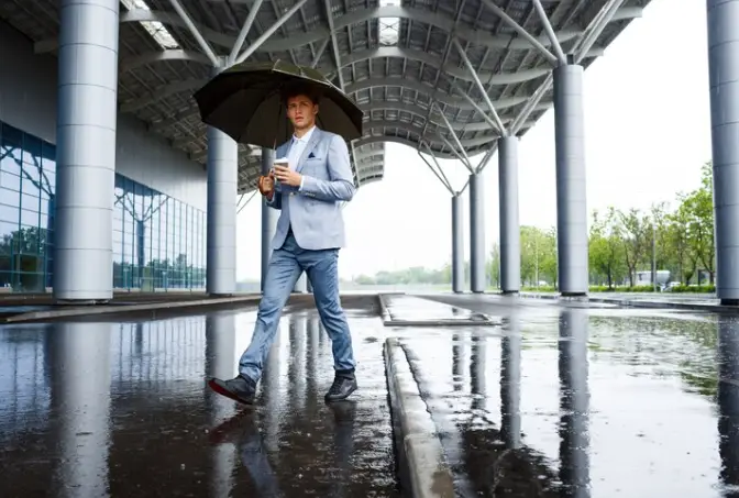 Man in blue suit walking with umbrella on wet pavement under modern architectural structure