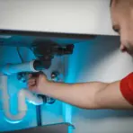 Person fixing white plumbing pipes under sink in brightly lit kitchen