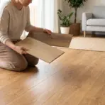 Person kneeling on wooden floor comparing laminate samples in bright living room