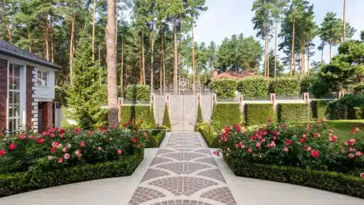 Brick pathway leading to ornate gate with lush garden and tall pine trees