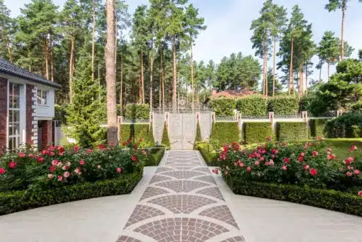 Brick pathway leading to ornate gate with lush garden and tall pine trees