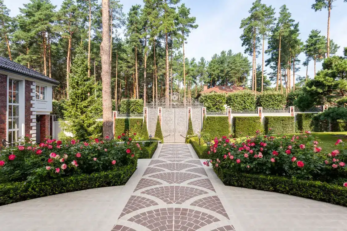 Brick pathway leading to ornate gate with lush garden and tall pine trees