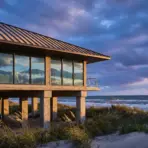 Modern beach house with large windows overlooking ocean under cloudy sky at sunset