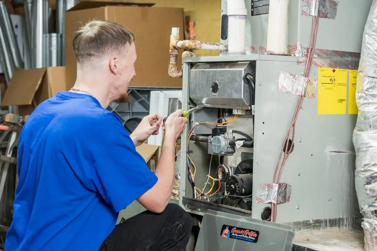 Technician in blue shirt repairing a furnace in a utility room with tools nearby