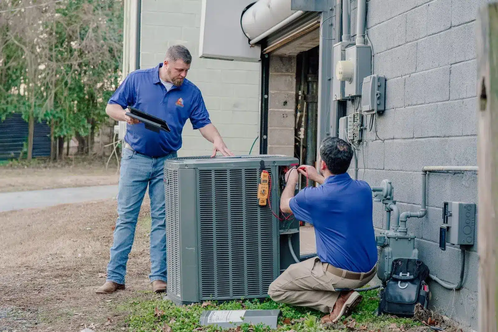 Two technicians working on an outdoor air conditioning unit next to a building wall