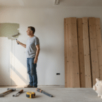 Man painting wall with paint roller in room with wooden planks and tools on floor