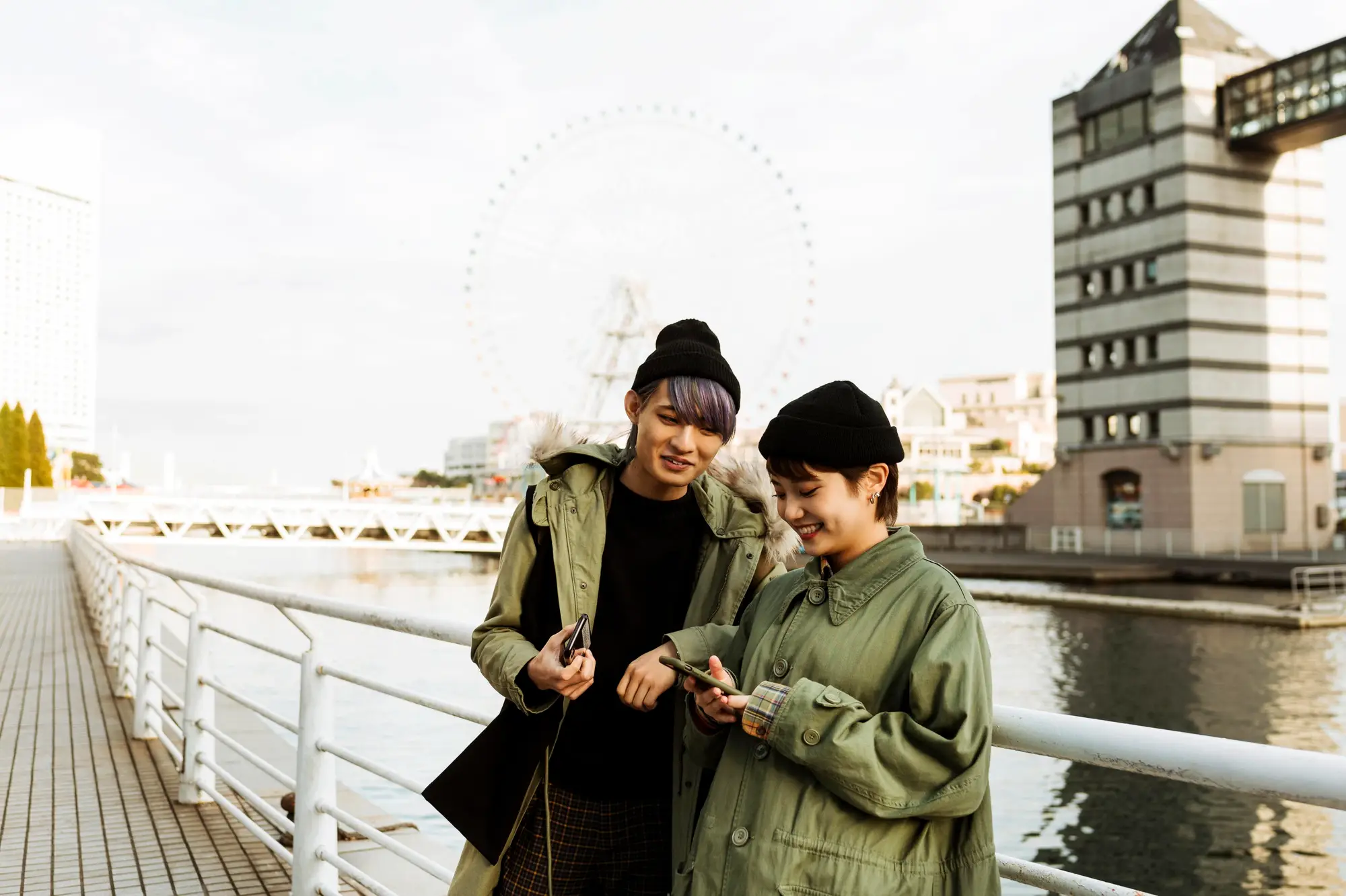Two people in green jackets smiling while looking at a phone outdoors by a waterfront.