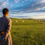 Man in traditional attire holding phone, standing in grassy field with yurts in distance