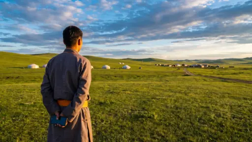 Man in traditional attire holding phone, standing in grassy field with yurts in distance