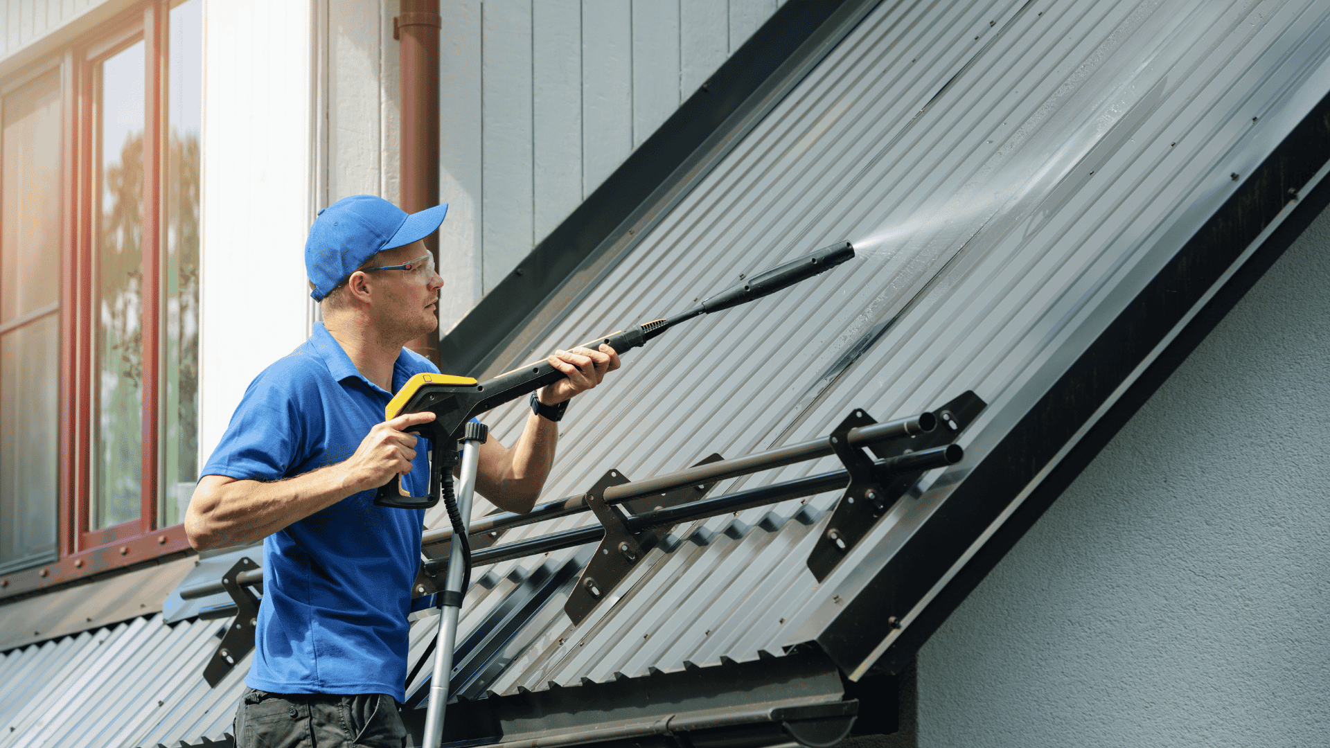 professional using a pressure washing wand to clean a sloped metal roof outside a home