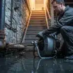 Worker using drain auger in flooded basement with stone walls and visible plumbing pipes