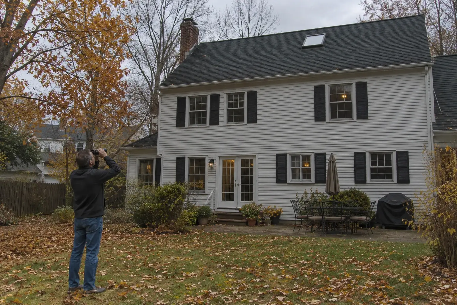 Person photographing a two-story house in a suburban yard with autumn leaves