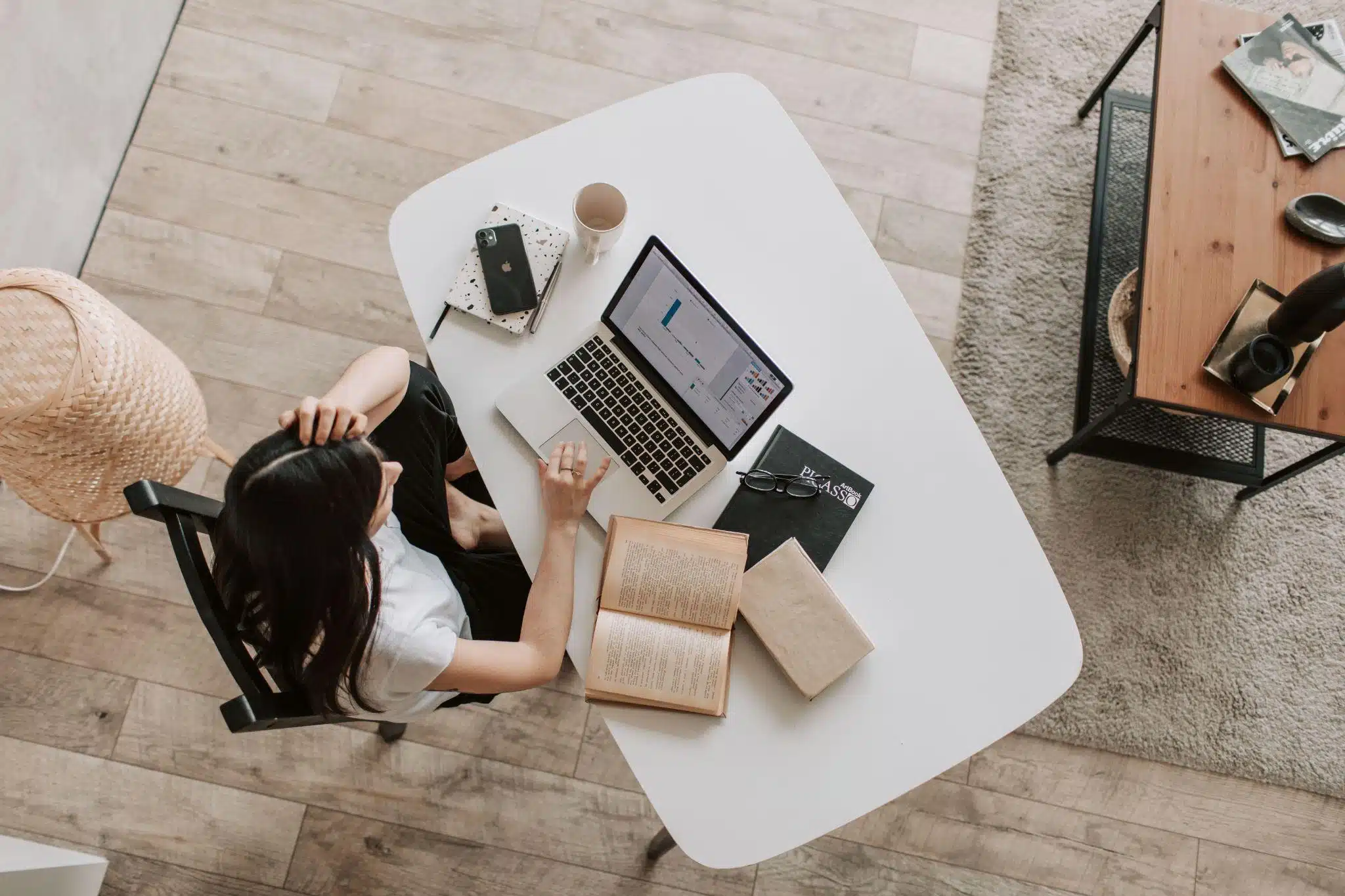 Person sitting at white desk with laptop, books, and smartphone in cozy indoor setting