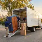 Movers loading a wooden cabinet and boxes into a truck on a tree-lined street