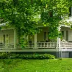 Victorian-style house porch surrounded by lush green trees and grass in a serene setting