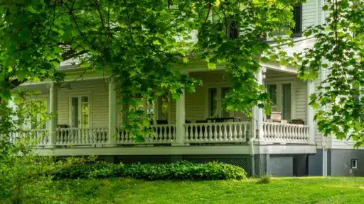 Victorian-style house porch surrounded by lush green trees and grass in a serene setting
