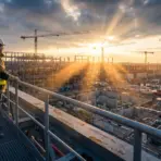 Construction worker in safety gear observing large construction site at sunrise with cranes in background