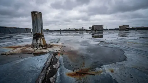 Rusty vent pipe and puddles on flat industrial rooftop under cloudy sky